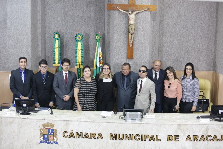 Vereador Lucas Aribé em pé rodeado dos componentes da mesa diretiva e vereadores presentes à Sessão Especial pelo Dia Internacional do Consumidor