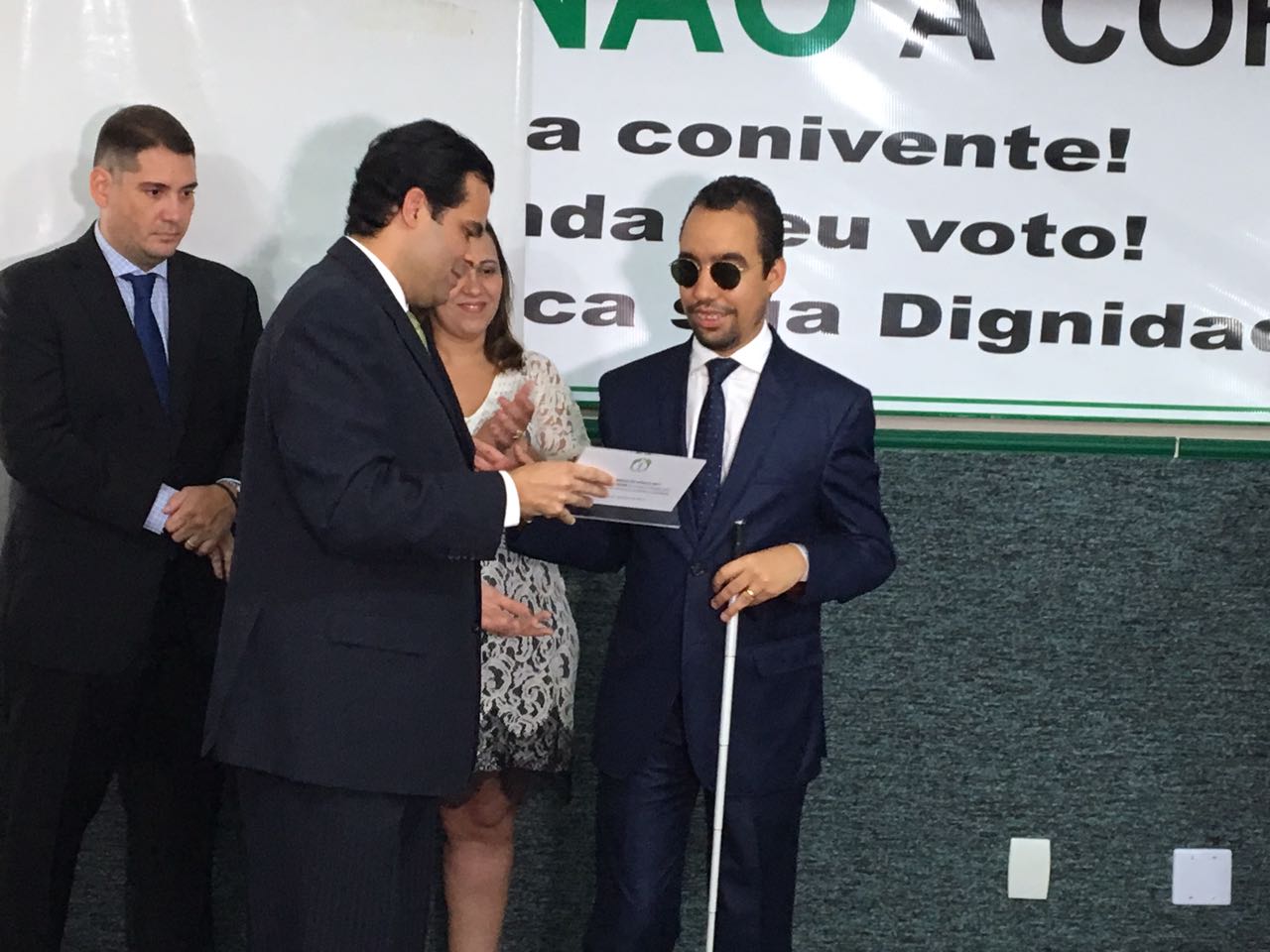 #PracegoVer: Lucas Aribé está segurando sua bengala com a mão esquerda enquanto recebe, com a mão direita, o troféu feito em acrílico, com mensagem do prêmio Amigo do Médico em Braile