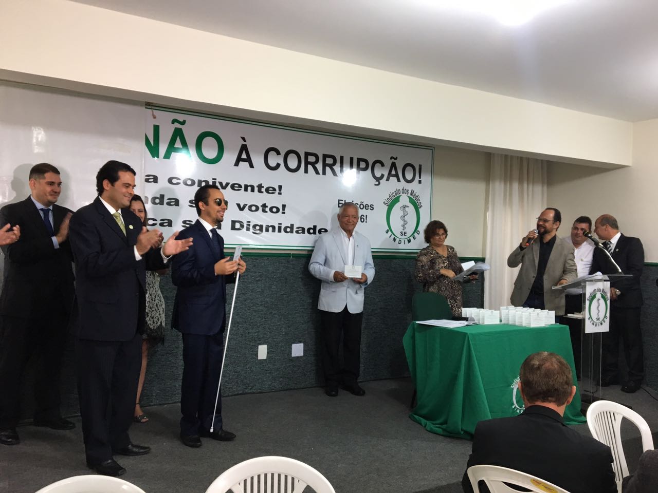 #PracegoVer: Lucas Aribé está à esquerda da foto, lendo a mensagem em Braile do prêmio, enquanto que na outra extremidade da imagem está o mestre de cerimônia próximo ao púlpito do auditório