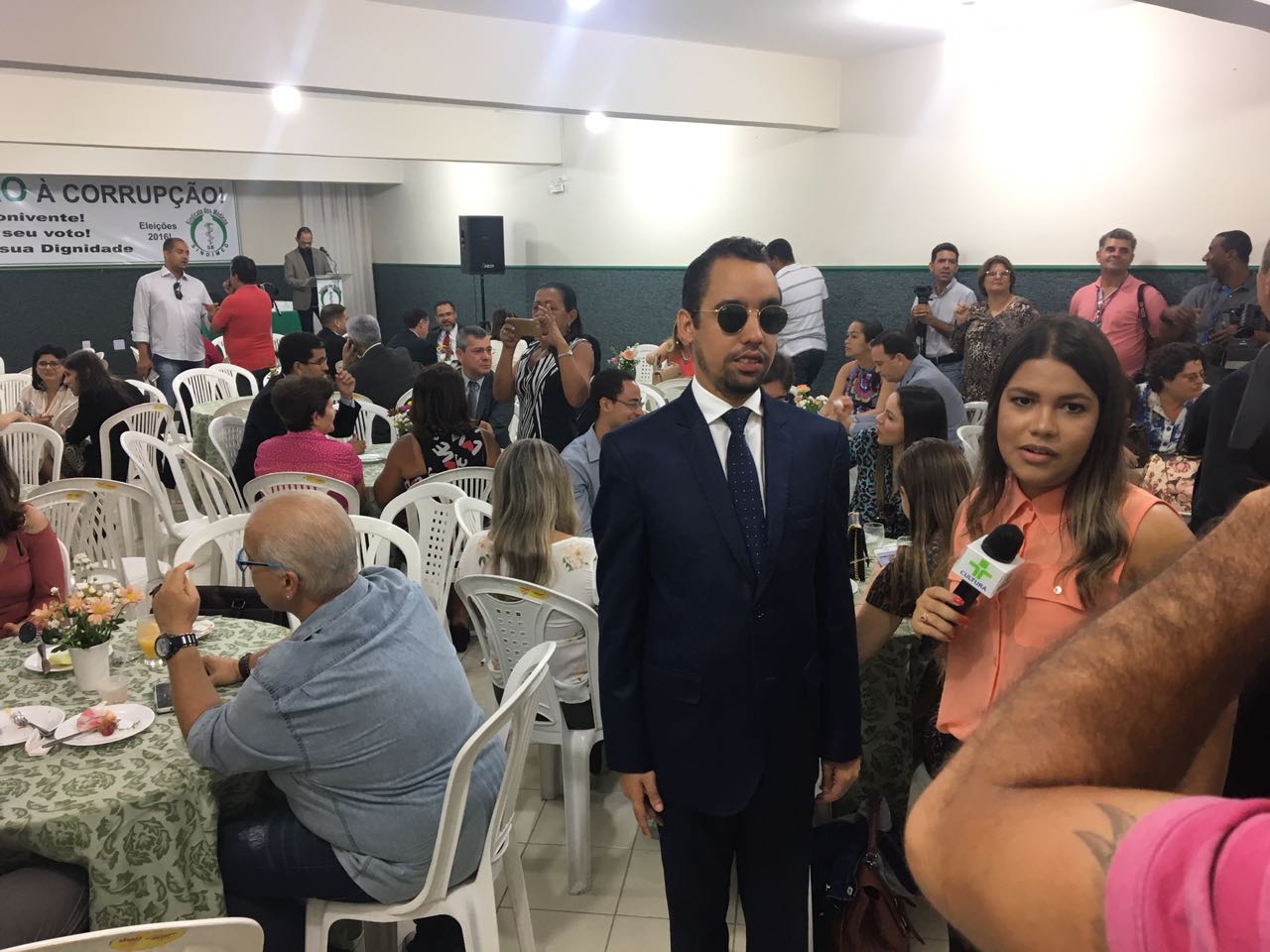#PraCegoVer: Lucas Aribé está à direita da foto sendo entrevistado pela equipe da TV Aperipê. Ao fundo, tem mesas com pessoas sentadas dentro do auditório. No final, dá para ver o palco