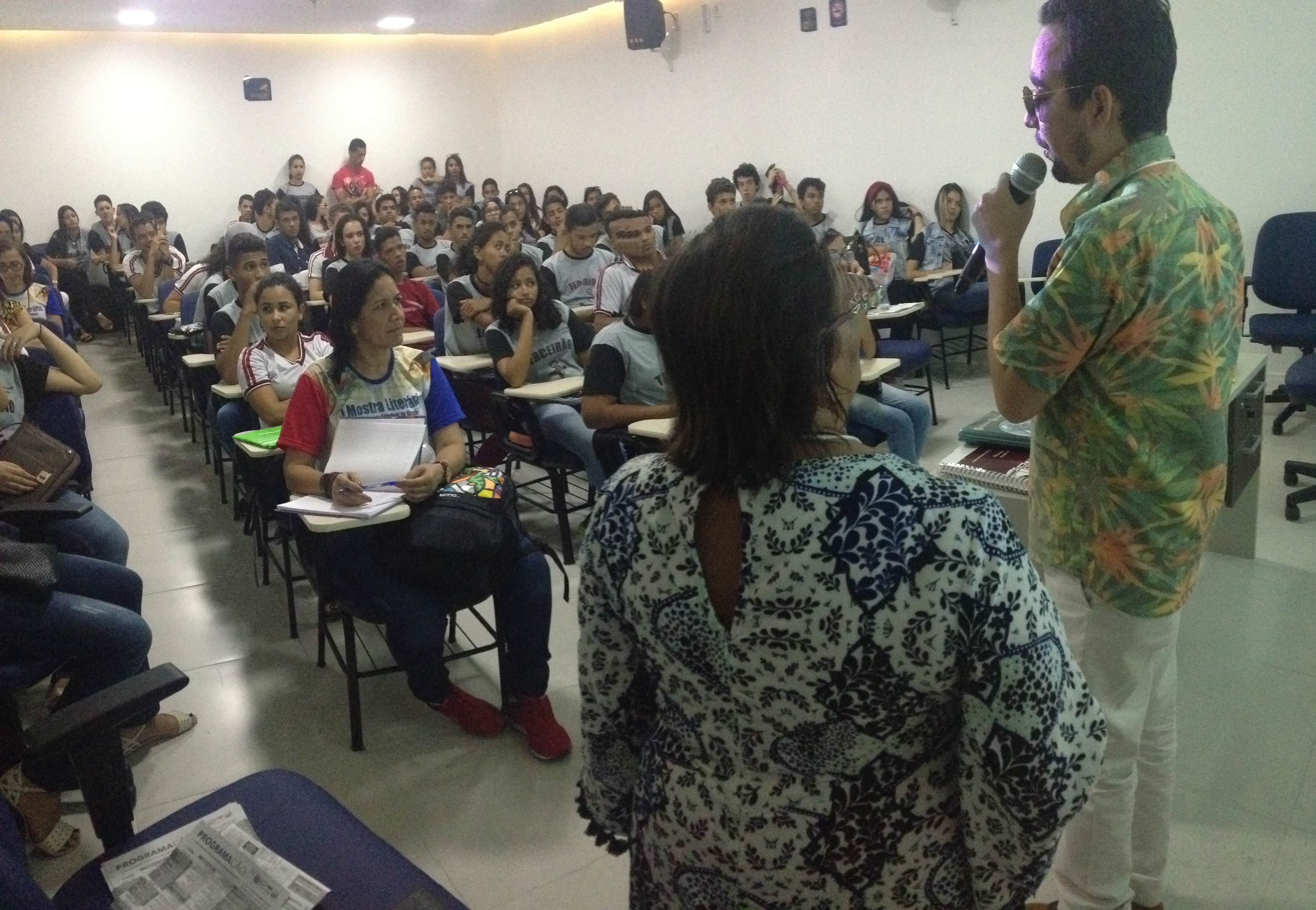 #PraCegoVer: Lucas e professora Margarida estão de costas na foto e alunos lotam auditório durante palestra