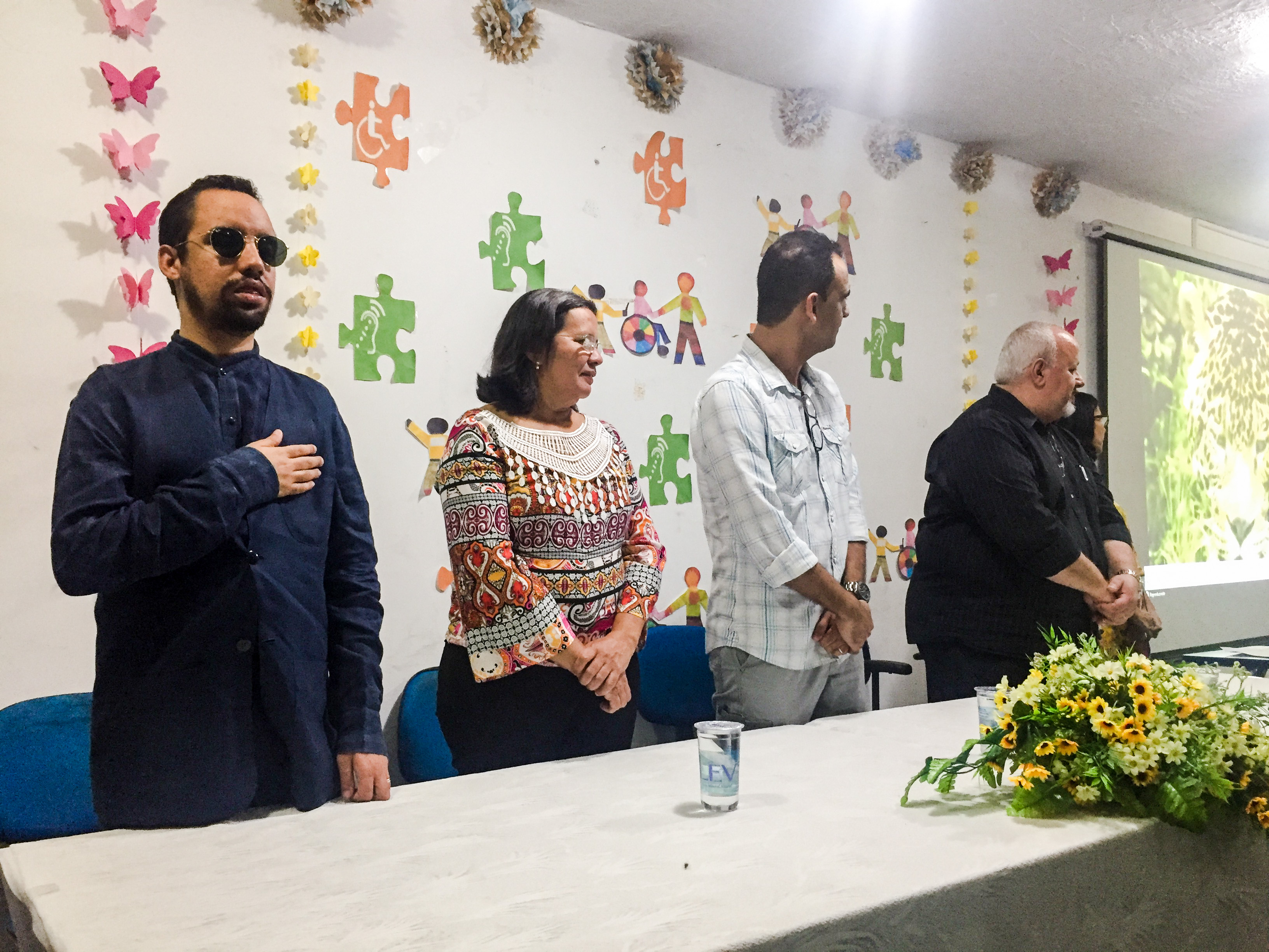 #PraCegoVer: O vereador Lucas Aribé está de pé fazendo parte da composição da mesa do evento. Está direita encostada no peito esquerdo atendo a execução do Hino Nacional 