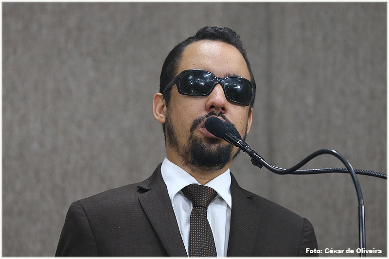 #PraCegoVer: O vereador Lucas Aribé está utilizando a tribuna da Câmara para se pronunciar. Ele está usando um terno marrom, camisa branca e gravata marrom com detalhes