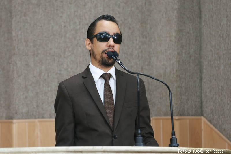 #PraCegoVer: O vereadro Lucas Aribé está de pé com um terno marrom, camisa branca e gravata marrom com detalhes falando na tribuna da Câmara 