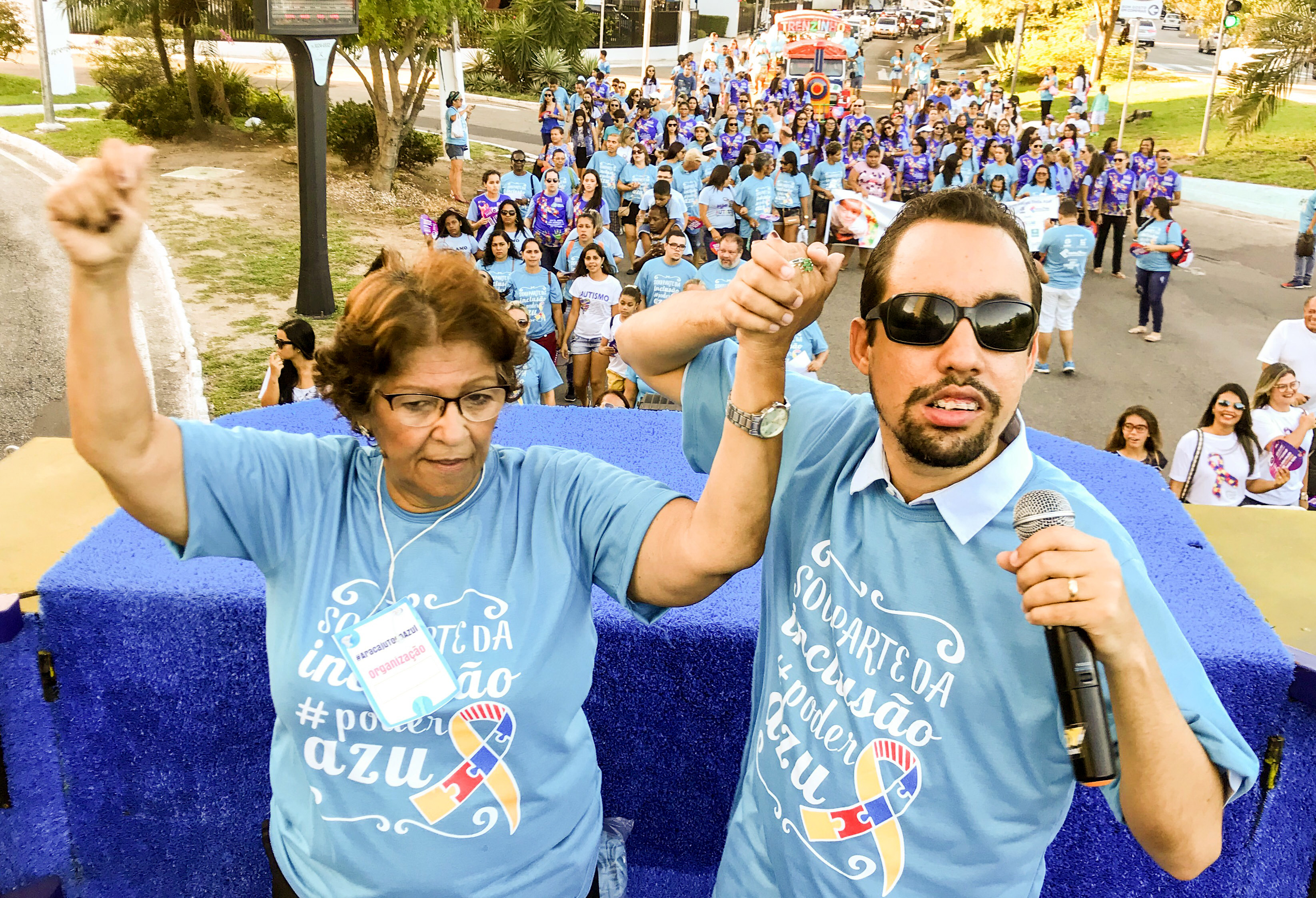 #PraCegoVer - Em cima do carro de som, de mãos dadas, a professora Maria do Carmo e Lucas Aribé conduzem uma legião de autistas e familiares
