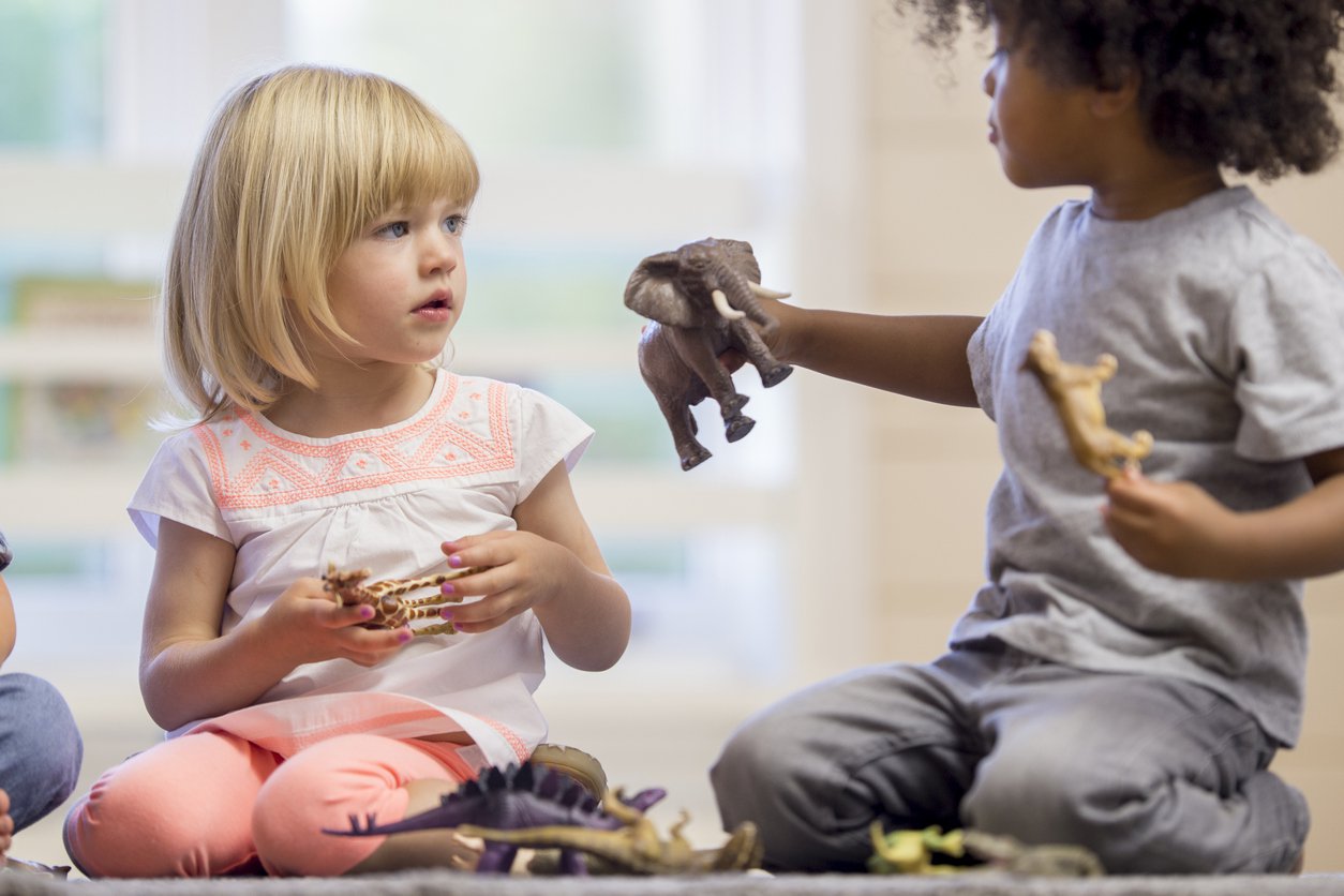 #PraCegoVer - Duas crianças brincam sentadas no chão, uma delas entrega um brinquedo à outra (Foto: Istock)