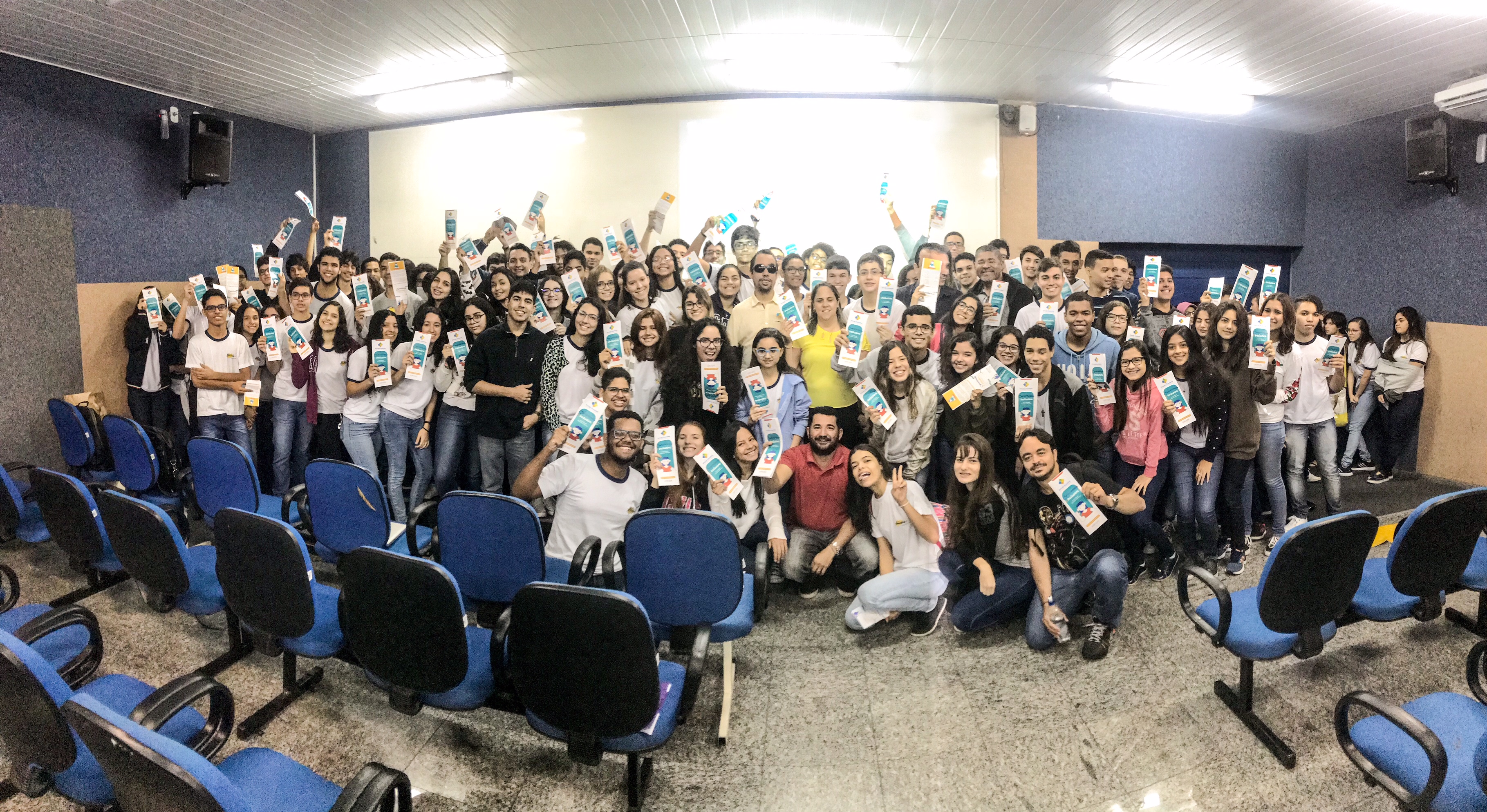#PraCegoVer: Lucas entre alunos e professores. Todos exibem a cartilha com dicas de comunicação inclusiva