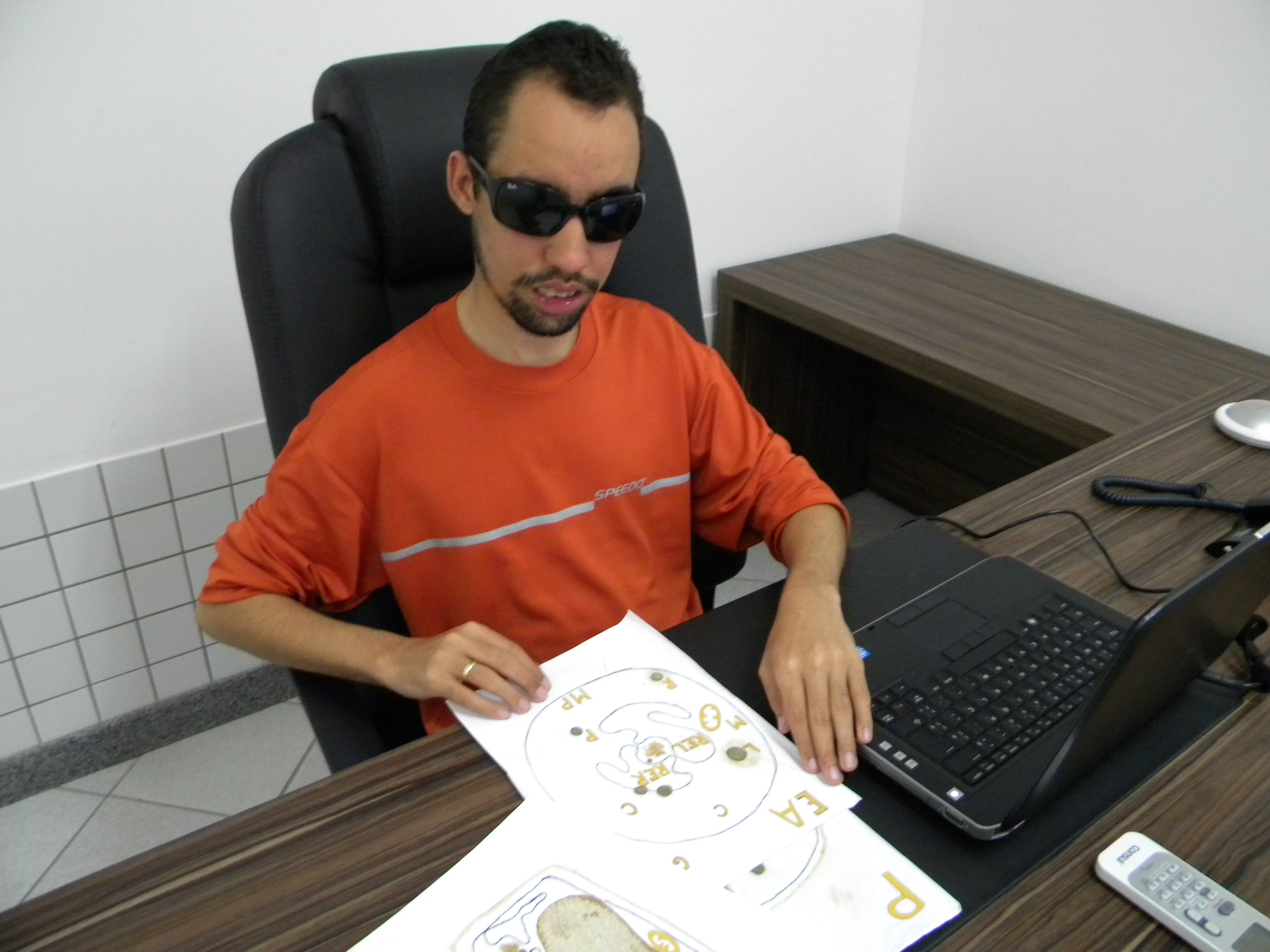 Lucas está sentado em frente ao notebook e está com os papéis dos desenhos das células nas mãos