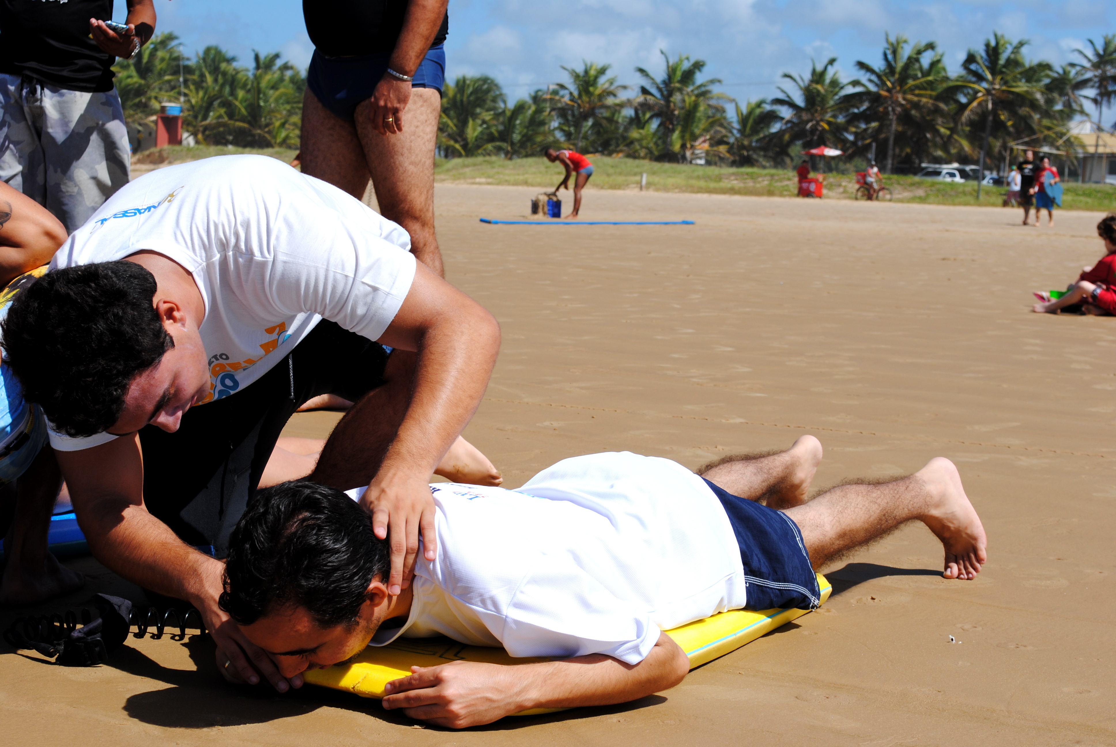 Lucas está deitado em um bodyboard na areia. Ao lado, o instrutor Lucas passa as dicas para Aribé