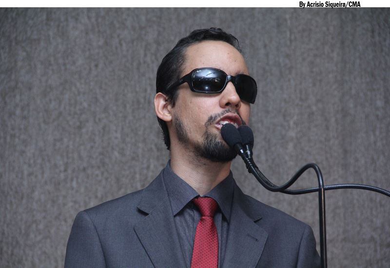 Usando terno de cor chumbo e gravata vermelha, Lucas está no centro da imagem. Ele ocupa a Tribuna da CMA
