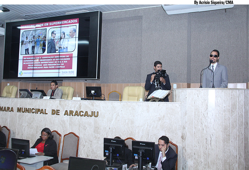Foto do plenário. Ao fundo, Lucas ocupa a Tribuna. O telão mostra imagens do Aracaju Acessível