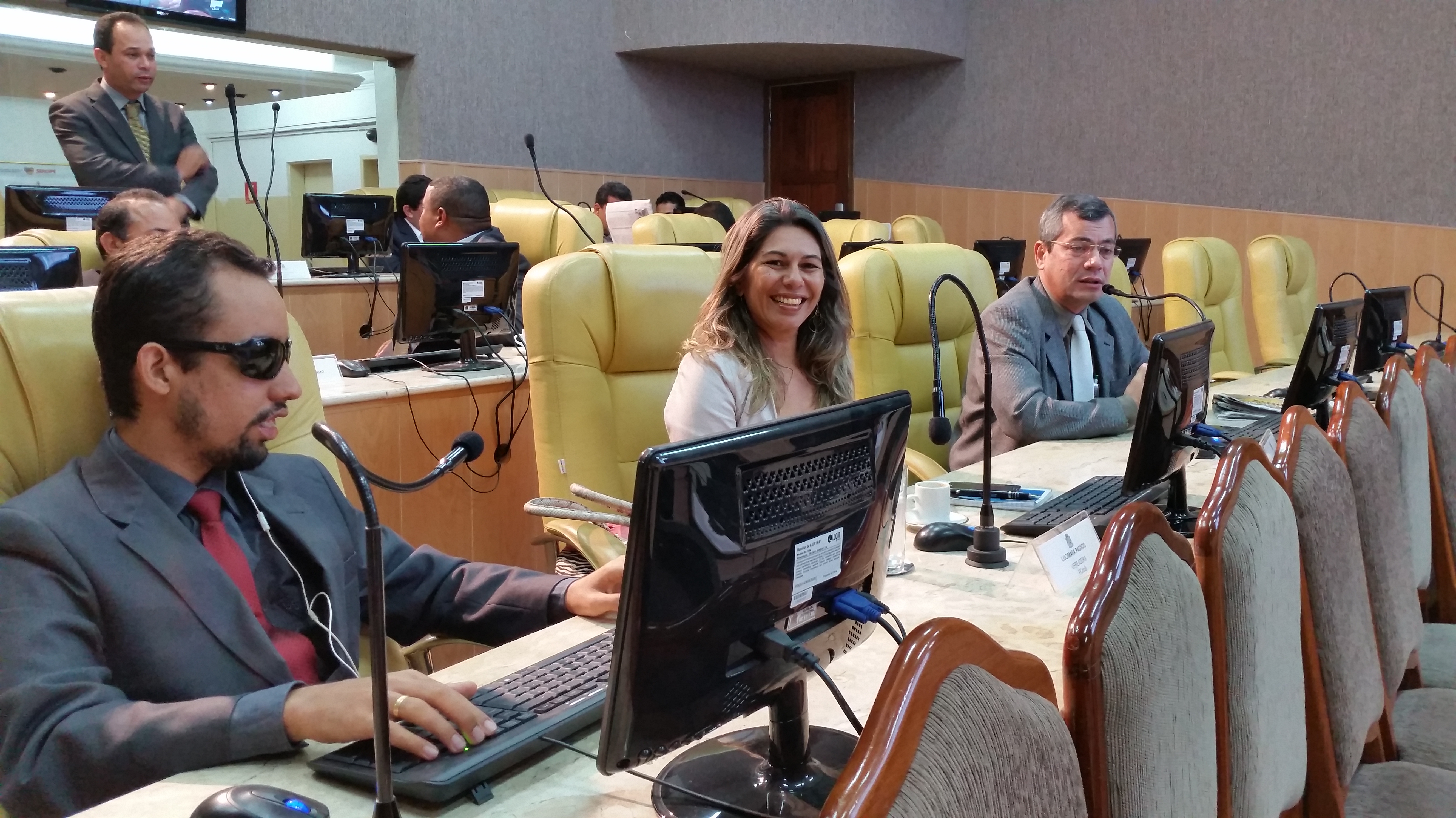 Lucas, em primeiro plano, está ao lado da vereadora Lucimara Passos. Ambos escutam o pronunciamento de Iran Barbosa, que elogia a iniciativa. Iran aparece no canto direito da imagem
