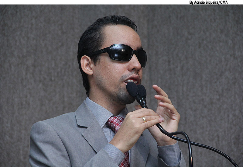 Na foto, Lucas segura o microfone da Tribuna com a mão direita. Ele veste terno cinza e blusa branca