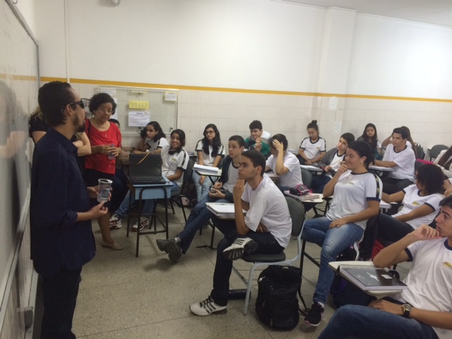 No canto esquerdo da foto, Lucas fala aos alunos em frente ao quadro branco. Estudantes estão sentados nas cadeiras