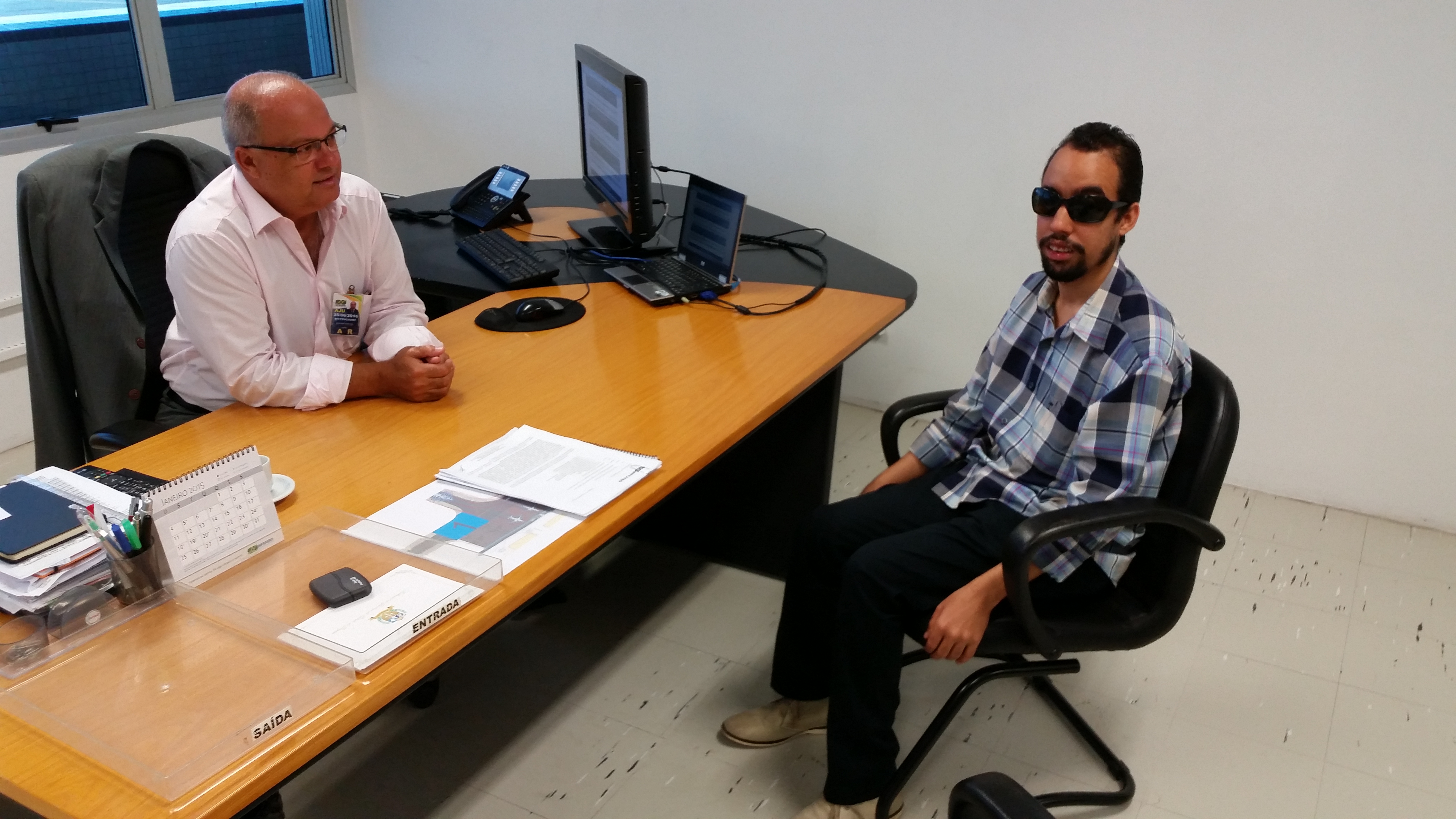 Imagem de Lucas e Luiz Alberto sentados. Entre eles, uma mesa de escritório